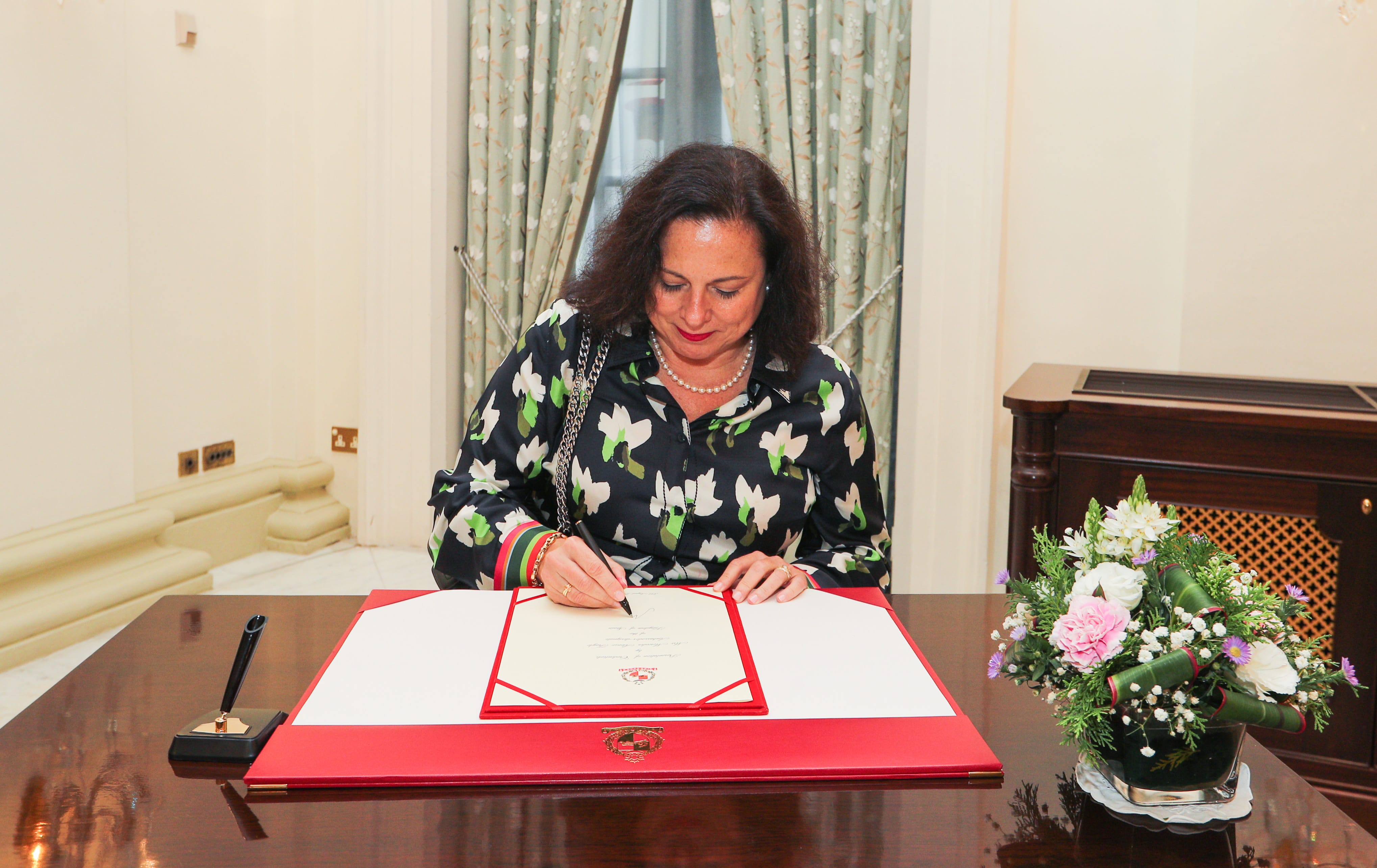 A woman signs a document with the Malta coat of arms, floral arrangement on desk.